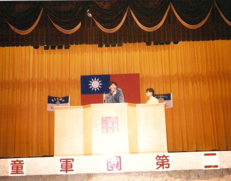 年輕TK在禮堂講台上持麥克風發言,背景懸掛中華民國國旗與童軍旗幟。